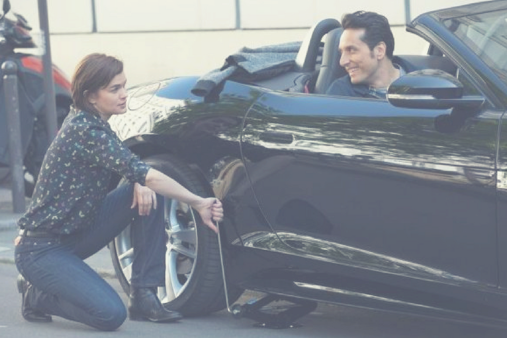 Casal. Ela troca o pneu, ele está dentro do carro.