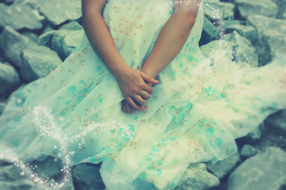 Menina sentada em chão de pedras, sozinha, com vestido que lembra os dos contos de fadas. Aparece apenas a metade inferior do seu corpo.